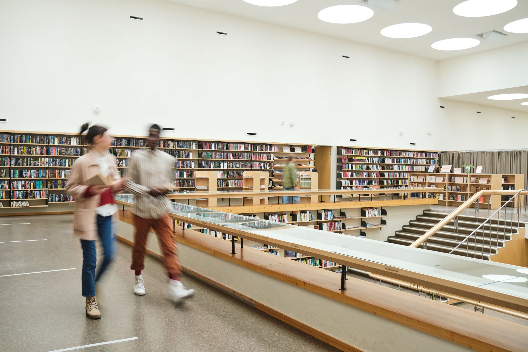 Library visitors walking past bookshelves on a sunlit upper level